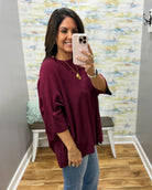 A woman takes a mirror selfie wearing a loose-fitting, deep burgundy short-sleeve top and light blue jeans in a home setting with a patterned wallpaper background.