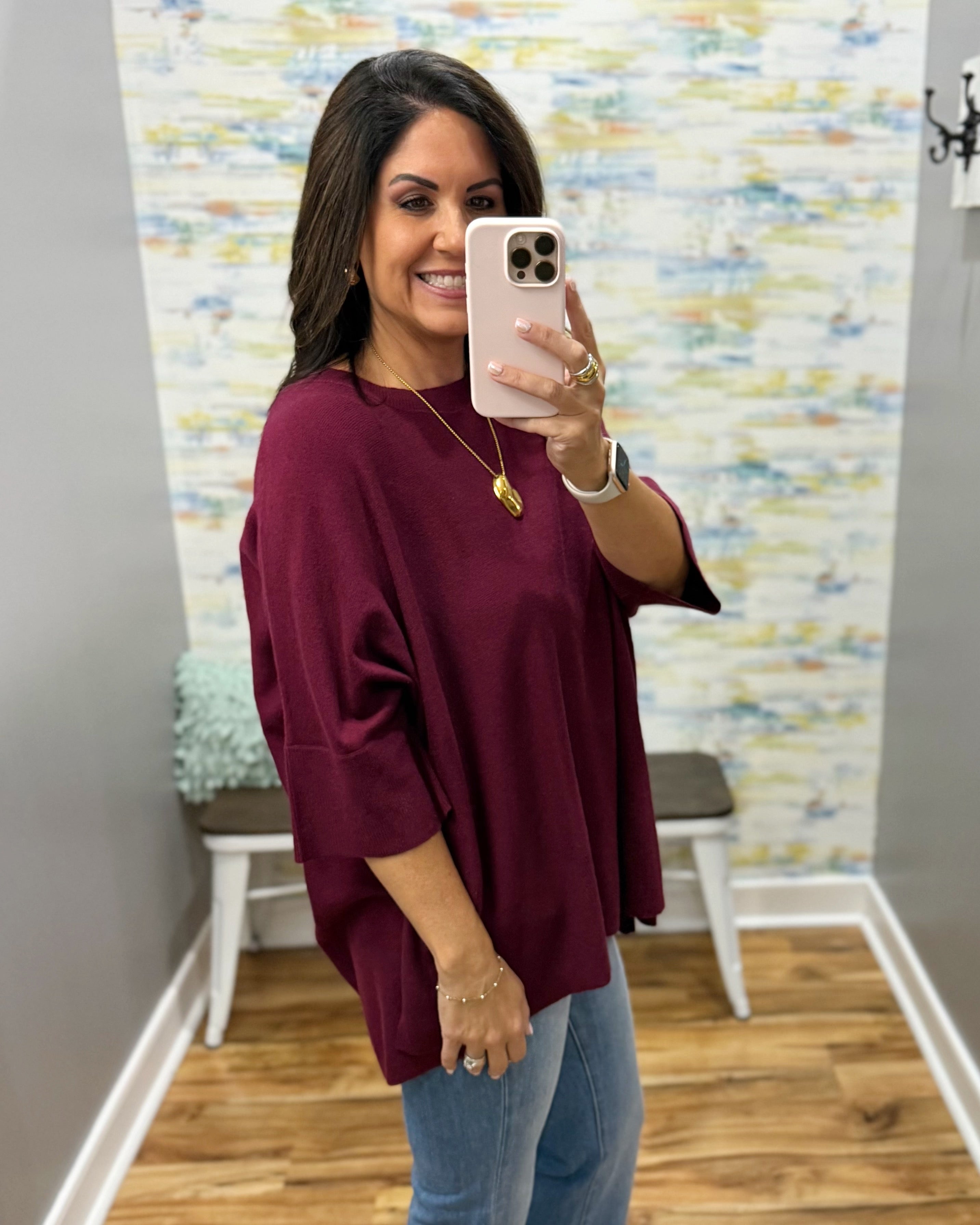 A woman takes a mirror selfie wearing a loose-fitting, deep burgundy short-sleeve top and light blue jeans in a home setting with a patterned wallpaper background.