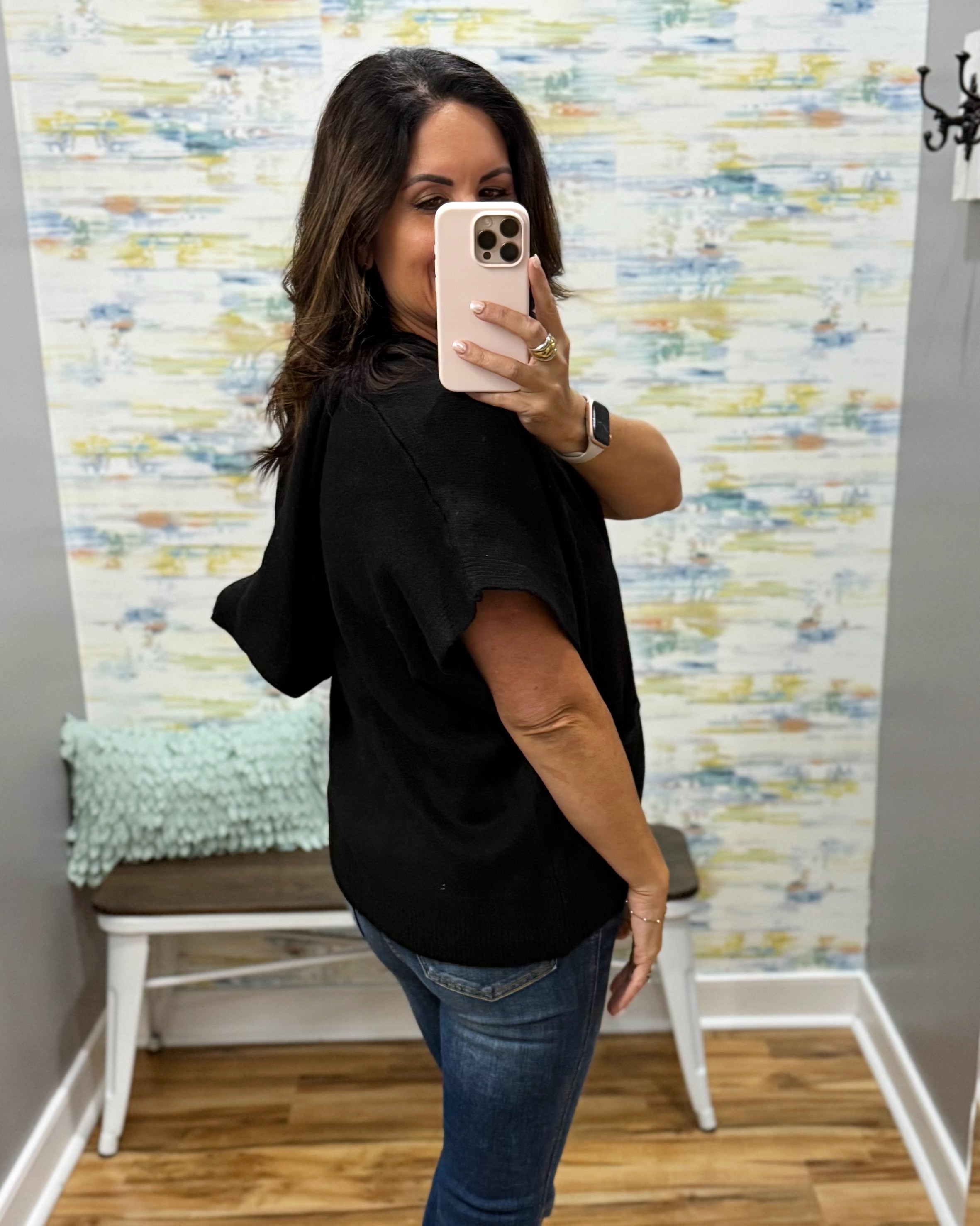 A woman takes a mirror selfie wearing a black short-sleeve top and blue jeans in front of a patterned wallpaper background.