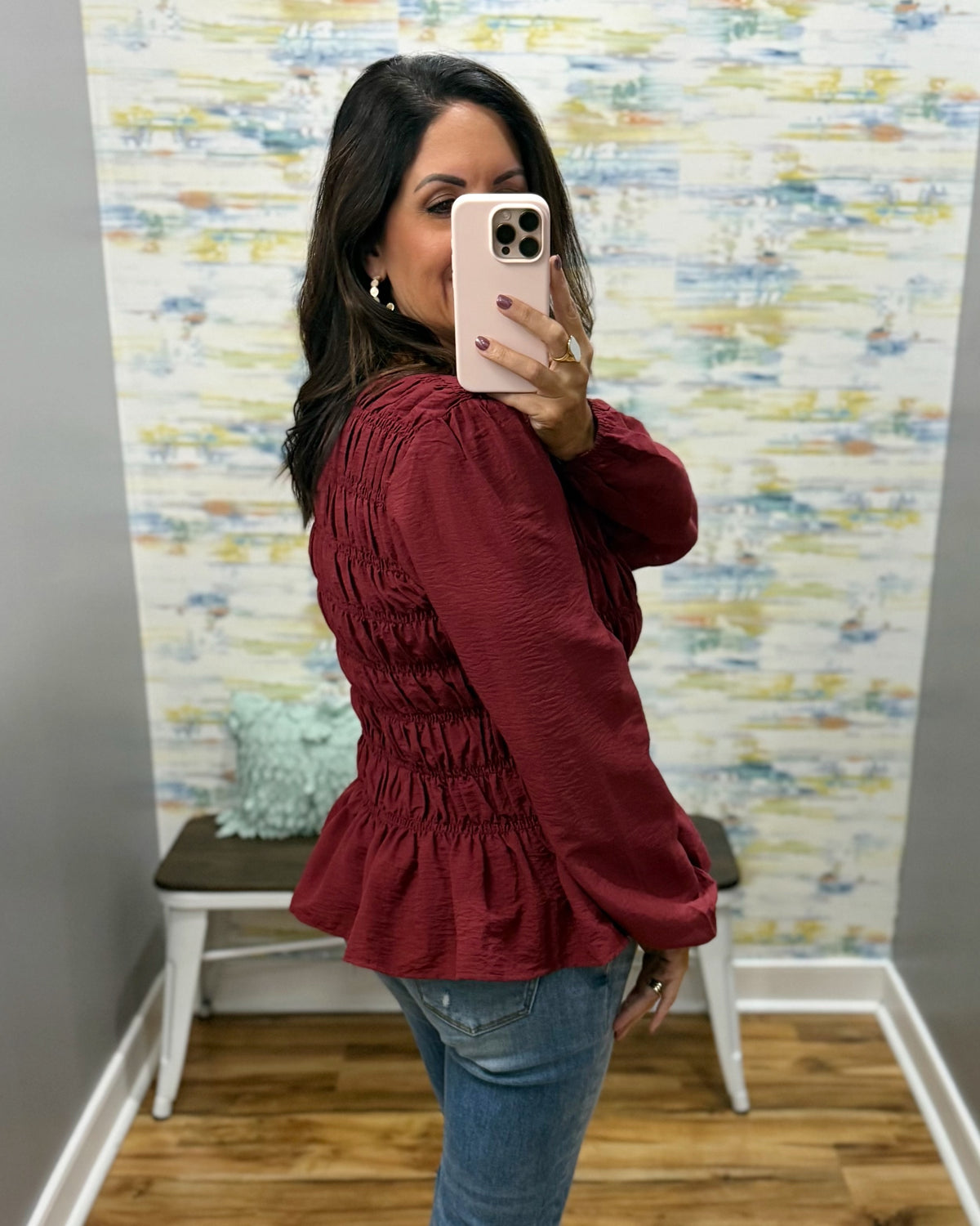 A woman takes a mirror selfie wearing a burgundy long-sleeve peplum blouse with a smocked bodice and blue jeans, standing in front of a patterned wallpaper background.