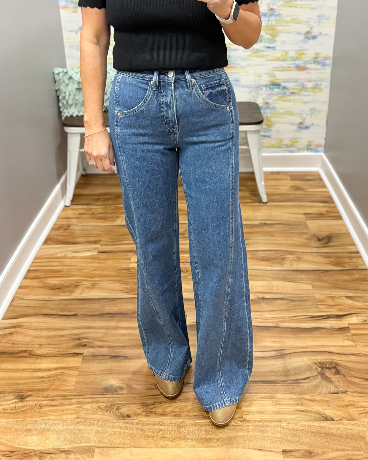 A person wearing mica wide-leg jeans, a black top, and tan pointed-toe heels, standing on a wood floor in an indoor room with a bench and abstract wall art in the background.