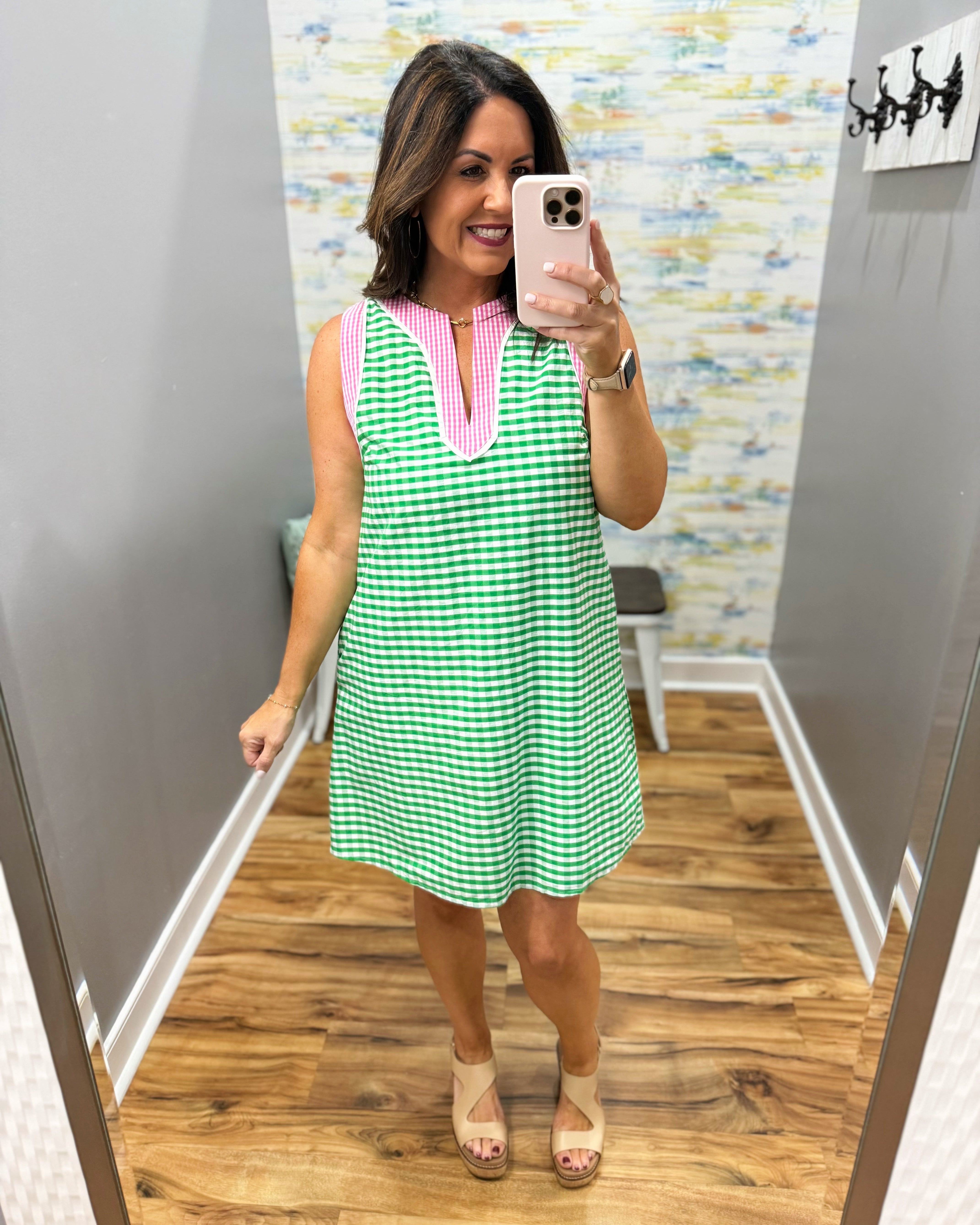 Woman taking a mirror selfie wearing a green and pink checkered dress in a room with a patterned wall.