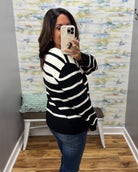 A woman takes a mirror selfie wearing a black and white horizontally striped knit sweater and blue jeans in a room with a patterned wallpaper background.