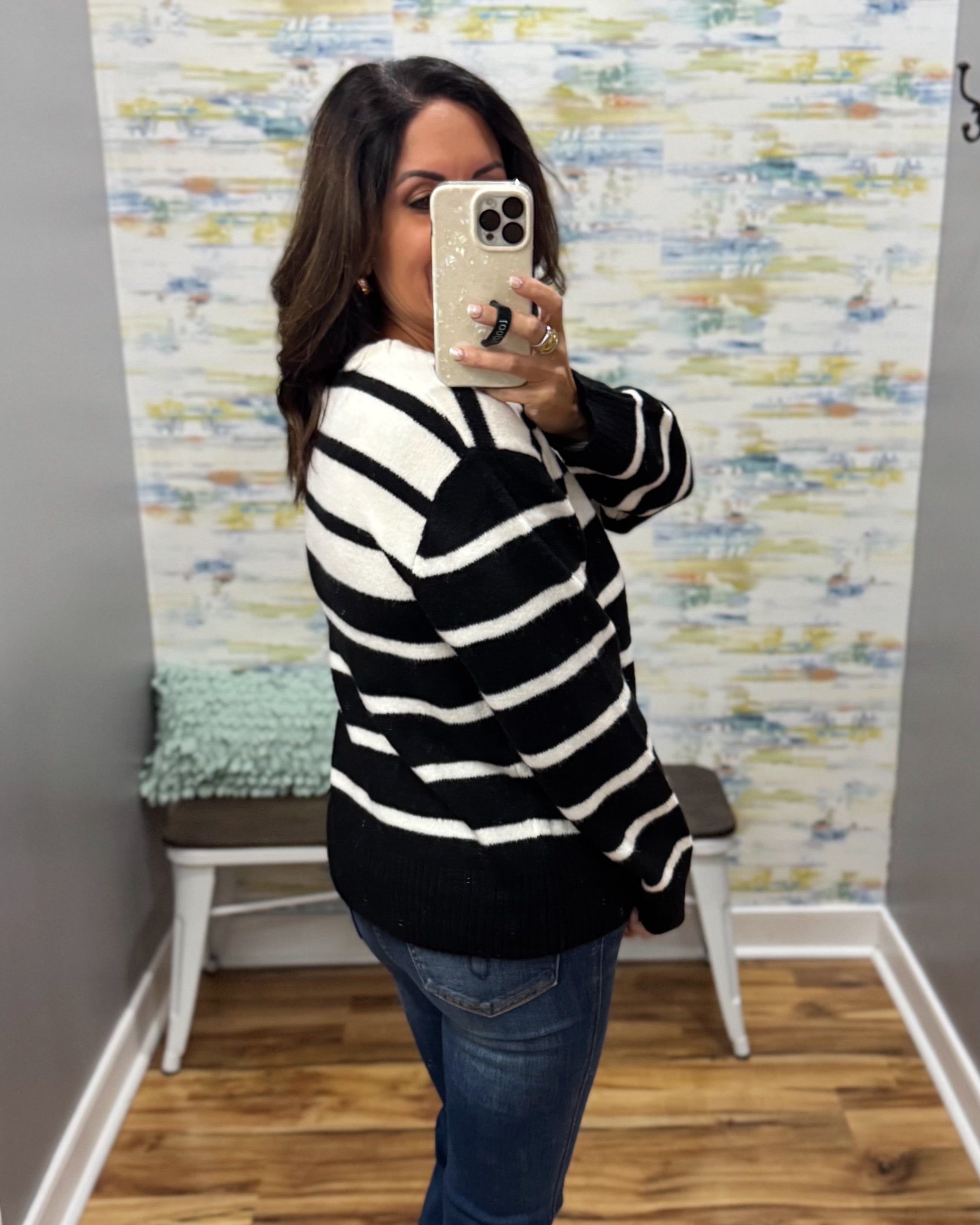 A woman takes a mirror selfie wearing a black and white horizontally striped knit sweater and blue jeans in a room with a patterned wallpaper background.