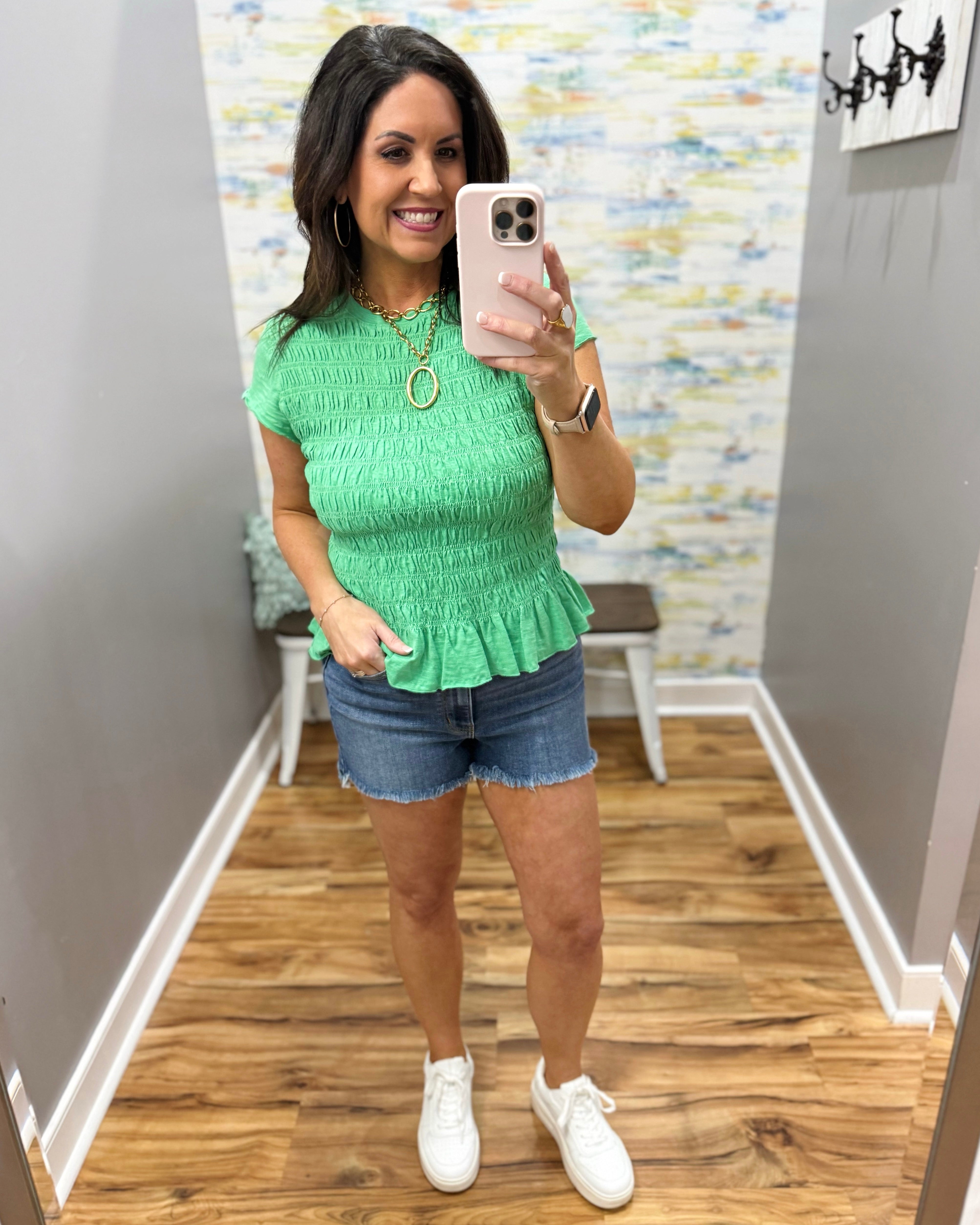 Woman wearing a green top and denim shorts in a room with a patterned wall.