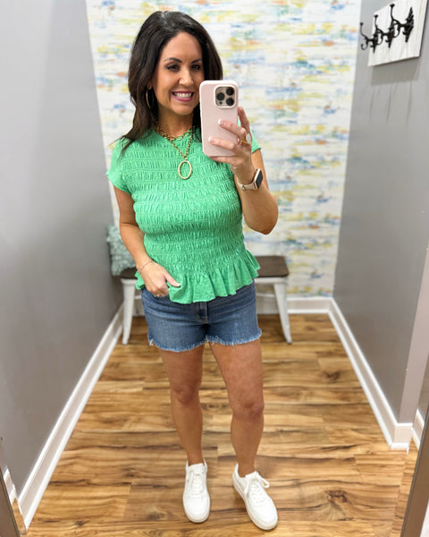 Woman wearing a green top and denim shorts in a room with a patterned wall.