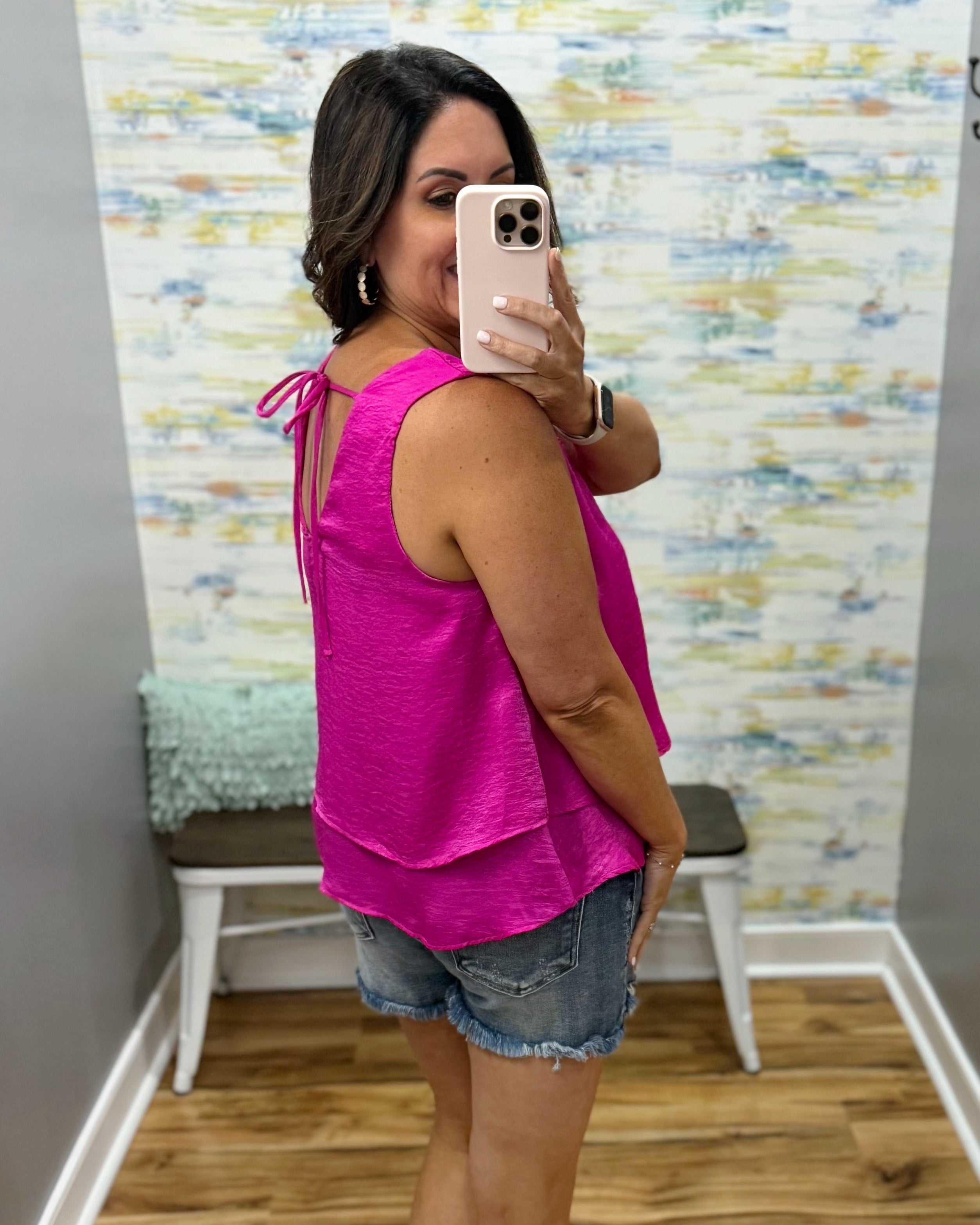 Woman taking a mirror selfie wearing a pink top and denim shorts in a room with a textured wall.