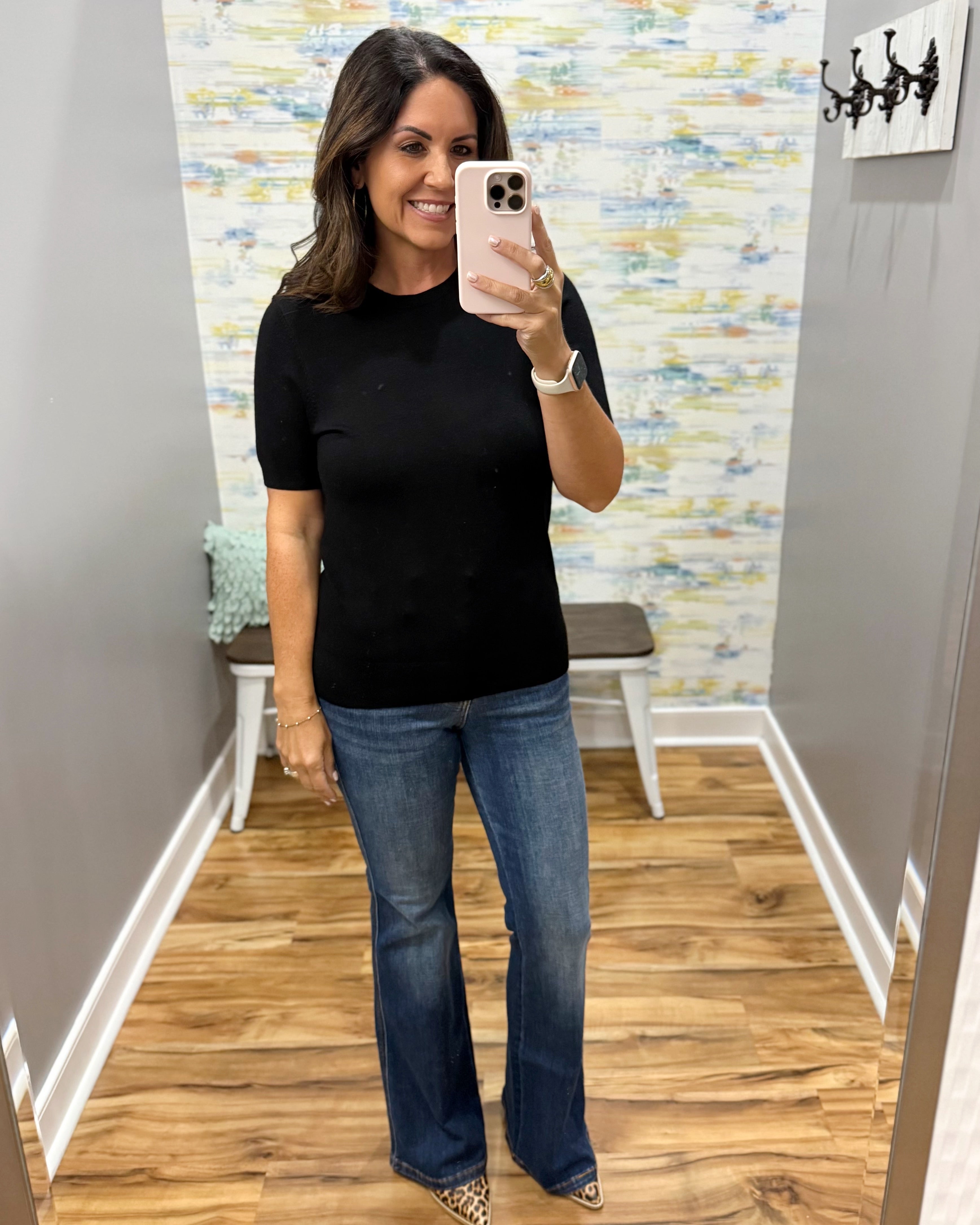 A woman takes a mirror selfie in a fitting room wearing a black short-sleeve crewneck top and blue flare jeans.