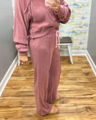 A person wearing a dusty rose pink two-piece lounge set consisting of a cropped long-sleeve top with snap buttons and matching wide-leg drawstring pants, standing on a wood floor in front of a mirror.