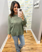 A woman takes a mirror selfie wearing a textured sage green long-sleeve knit top and light blue jeans in a home hallway.