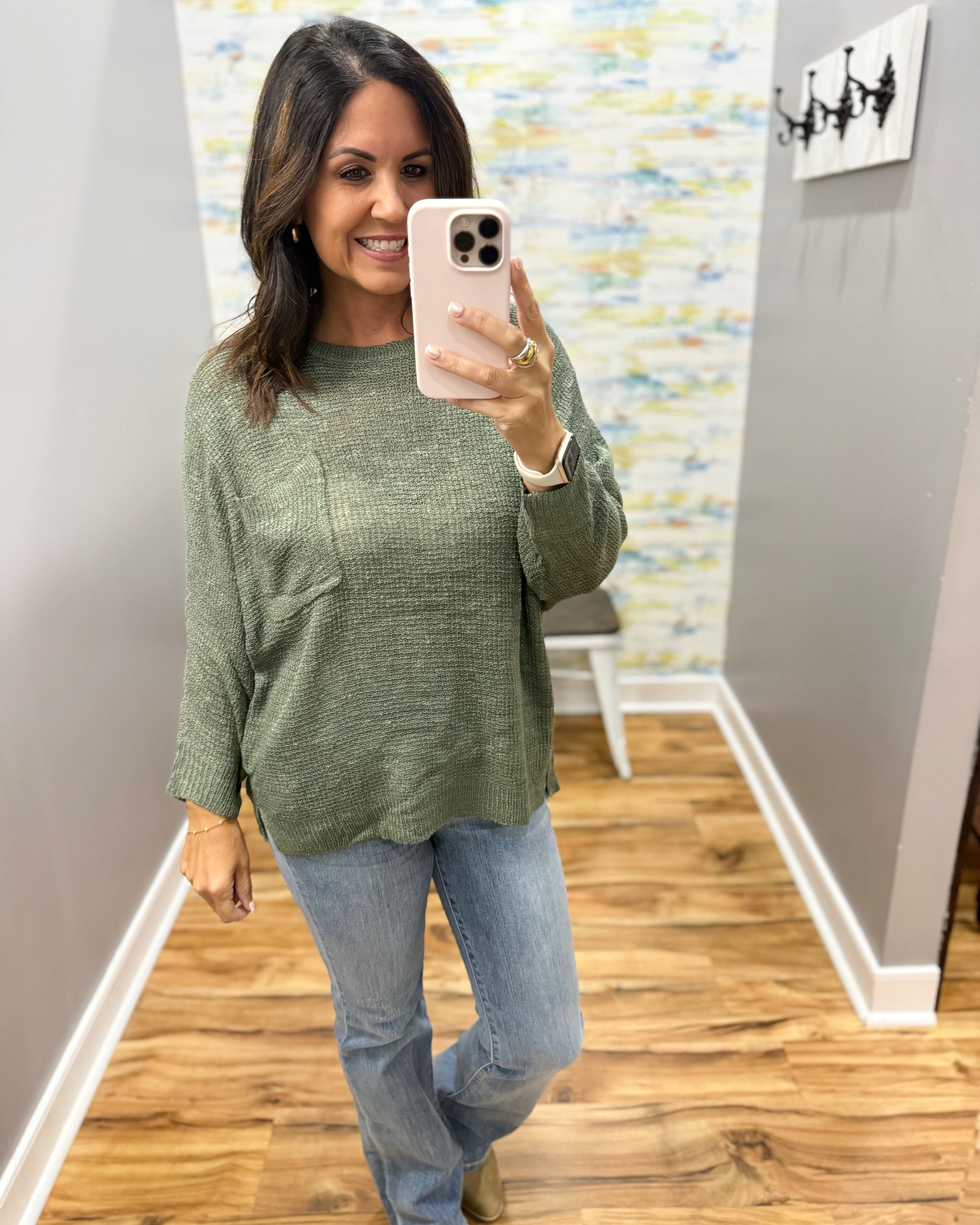 A woman takes a mirror selfie wearing a textured sage green long-sleeve knit top and light blue jeans in a home hallway.