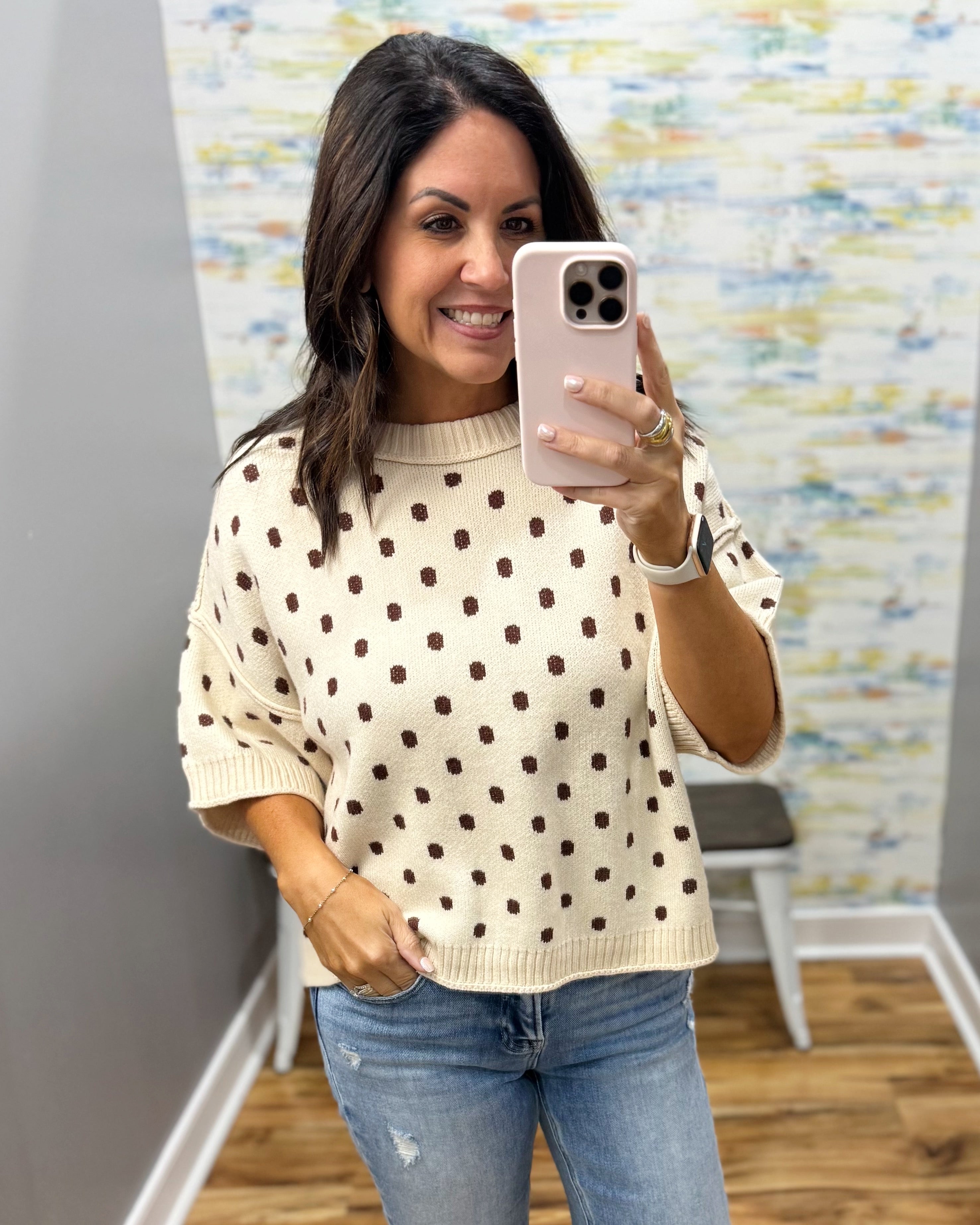 A woman takes a mirror selfie wearing a cream-colored knit sweater with brown polka dots and blue jeans in an indoor setting.