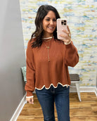 A woman takes a mirror selfie wearing a rust-orange knit sweater with a scalloped white hem and cuffs, paired with blue jeans and gold necklaces.