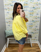 Woman taking a mirror selfie wearing a yellow sweater and denim shorts in a room with colorful wall art.