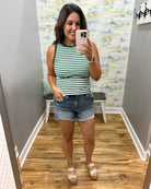 Woman taking a mirror selfie wearing a striped tank top and denim shorts in a room with a patterned wall.