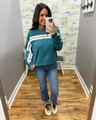 A woman takes a mirror selfie wearing a teal long-sleeve sweatshirt with white athletic stripes on the sleeves, paired with blue jeans and white sneakers.