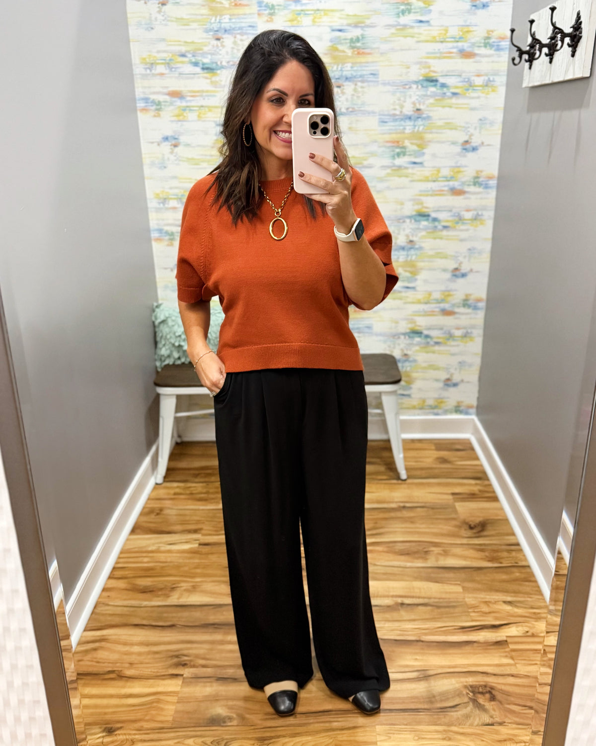 A woman takes a mirror selfie in a fitting room wearing a rust-orange short-sleeve knit top and black wide-leg pants.