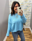 A woman takes a mirror selfie wearing a light blue knit sweater with a chest pocket and blue jeans in an indoor room with wood floors and a patterned wall.