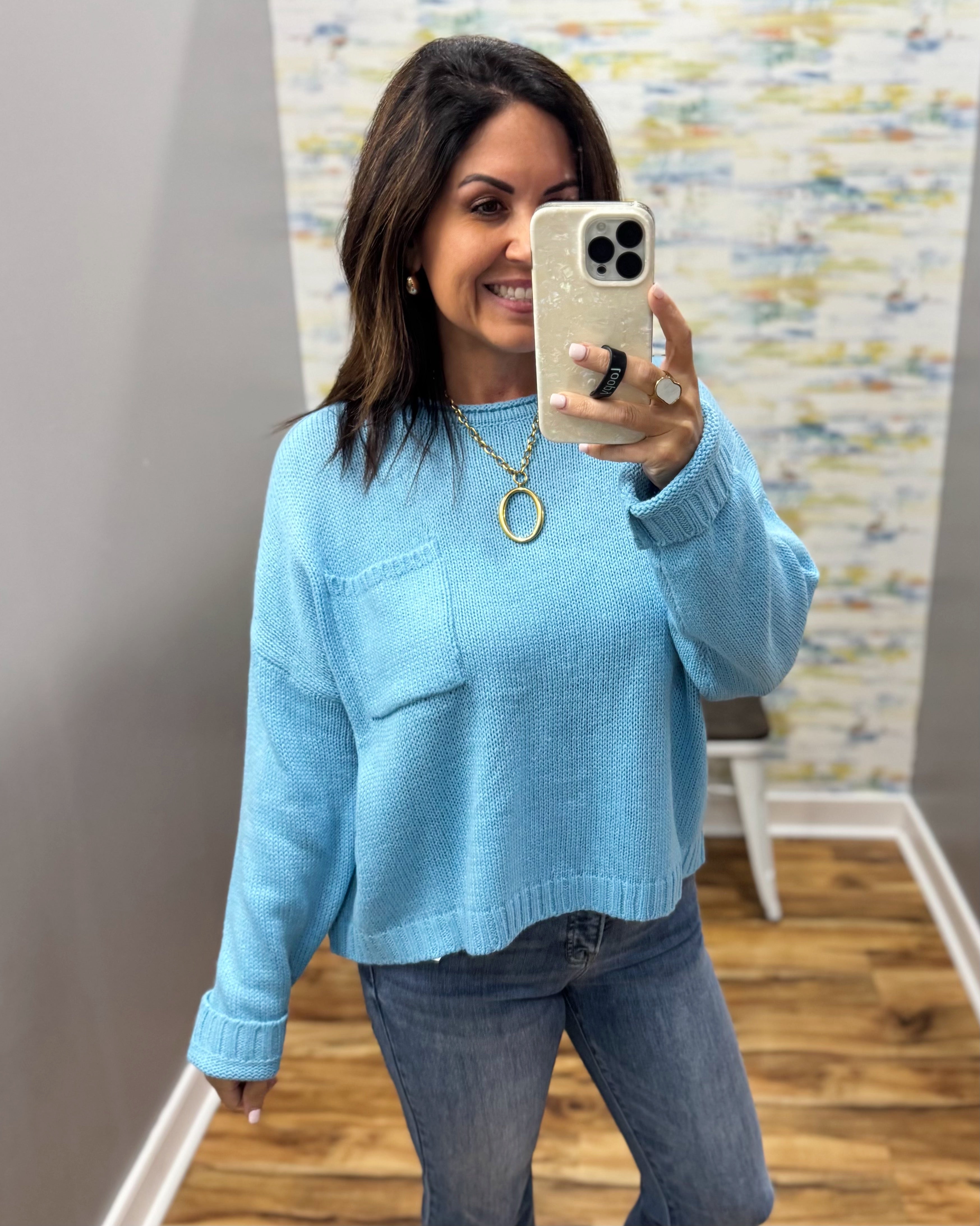 A woman takes a mirror selfie wearing a light blue knit sweater with a chest pocket and blue jeans in an indoor room with wood floors and a patterned wall.