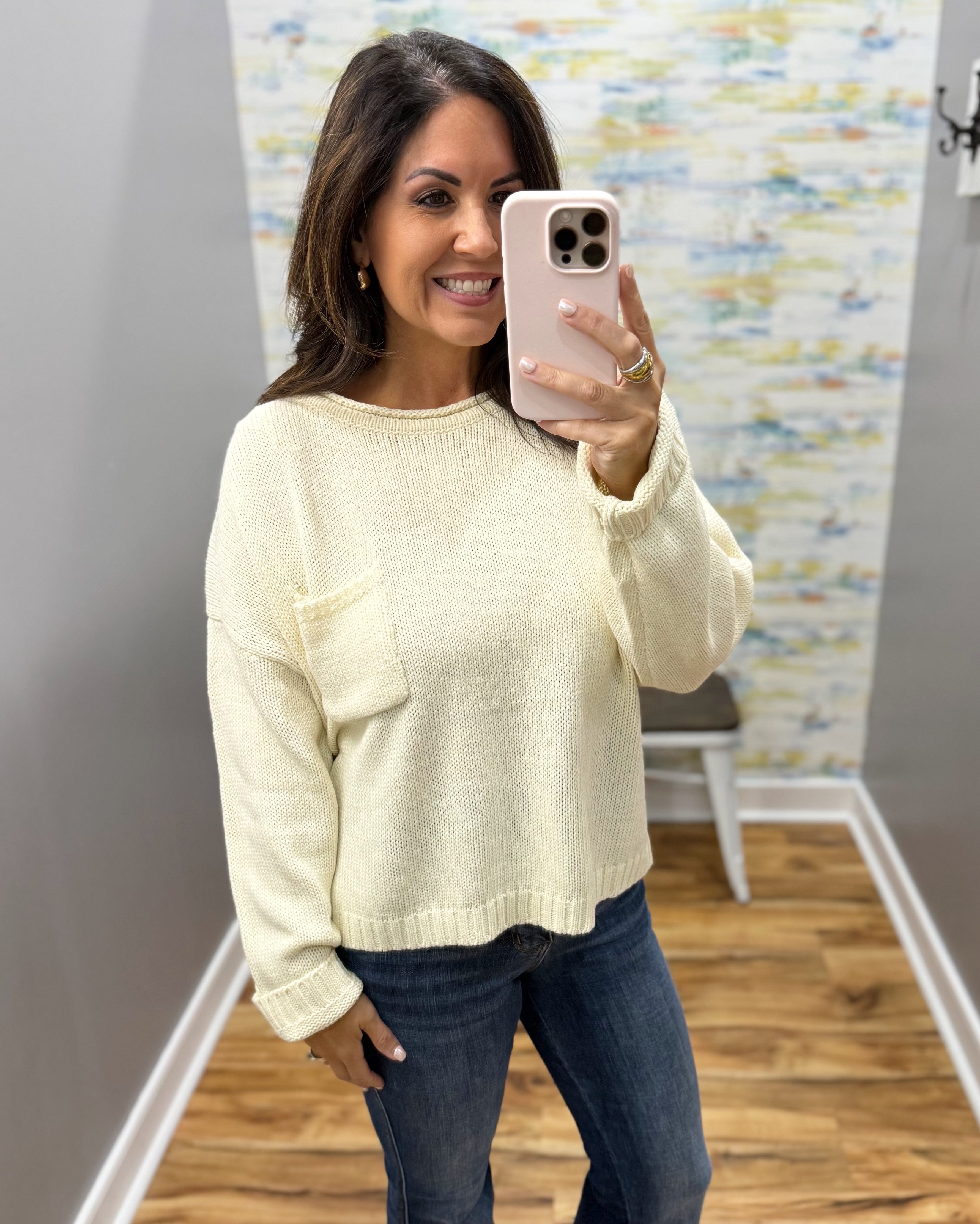 A woman takes a mirror selfie wearing a cream-colored knit sweater with a chest pocket and dark blue jeans, holding a light pink smartphone.