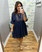 Woman taking a mirror selfie wearing a navy dress with colorful embroidery in a room with patterned wallpaper.