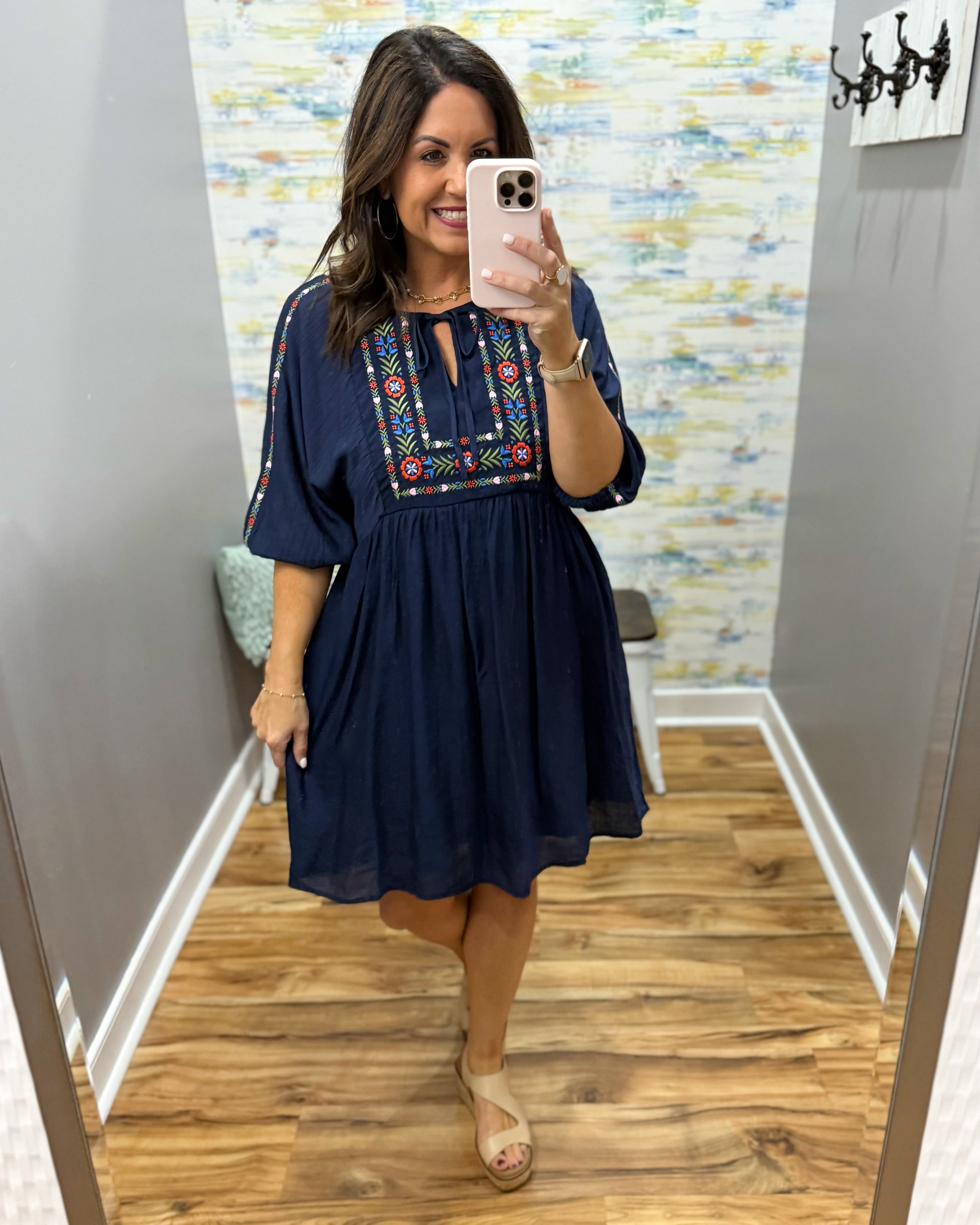 Woman taking a mirror selfie wearing a navy dress with colorful embroidery in a room with patterned wallpaper.