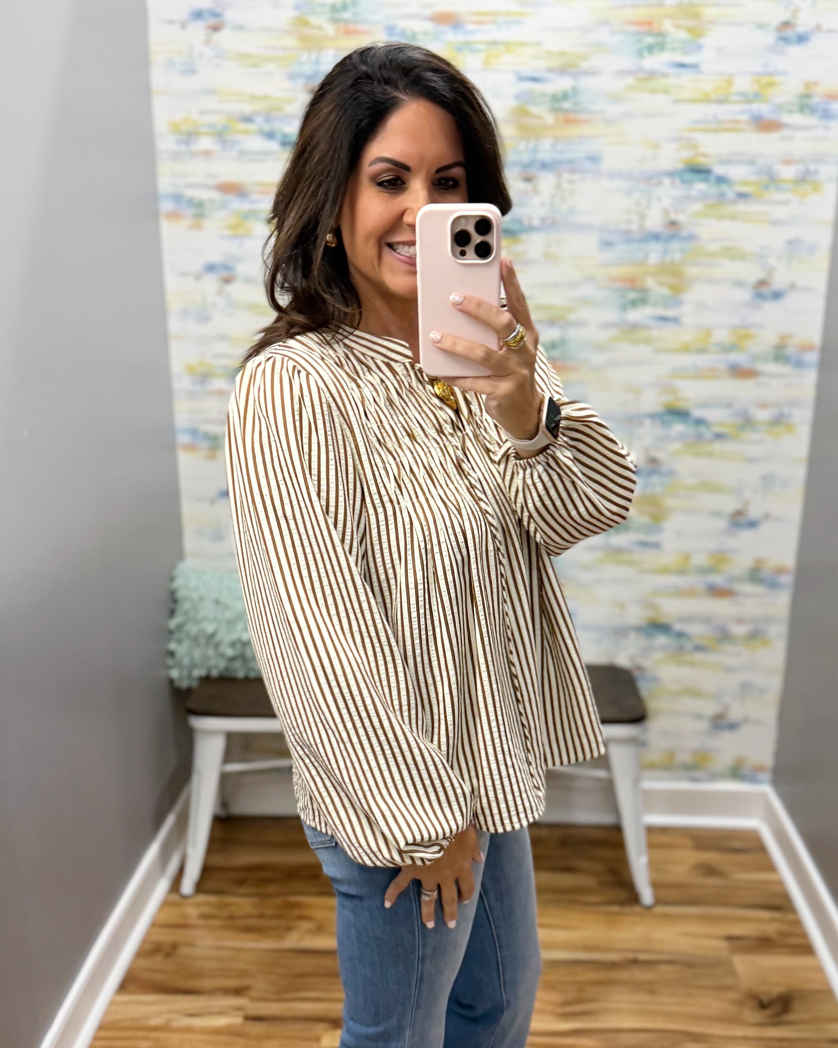 A woman takes a mirror selfie wearing a beige and white vertically striped long-sleeve blouse with a ruffled high neckline, paired with blue jeans.