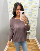A woman takes a mirror selfie wearing a textured, long-sleeve crewneck top in a dusty mauve-brown color, paired with blue jeans. The background shows a gray wall, a colorful abstract print, and a wooden floor.