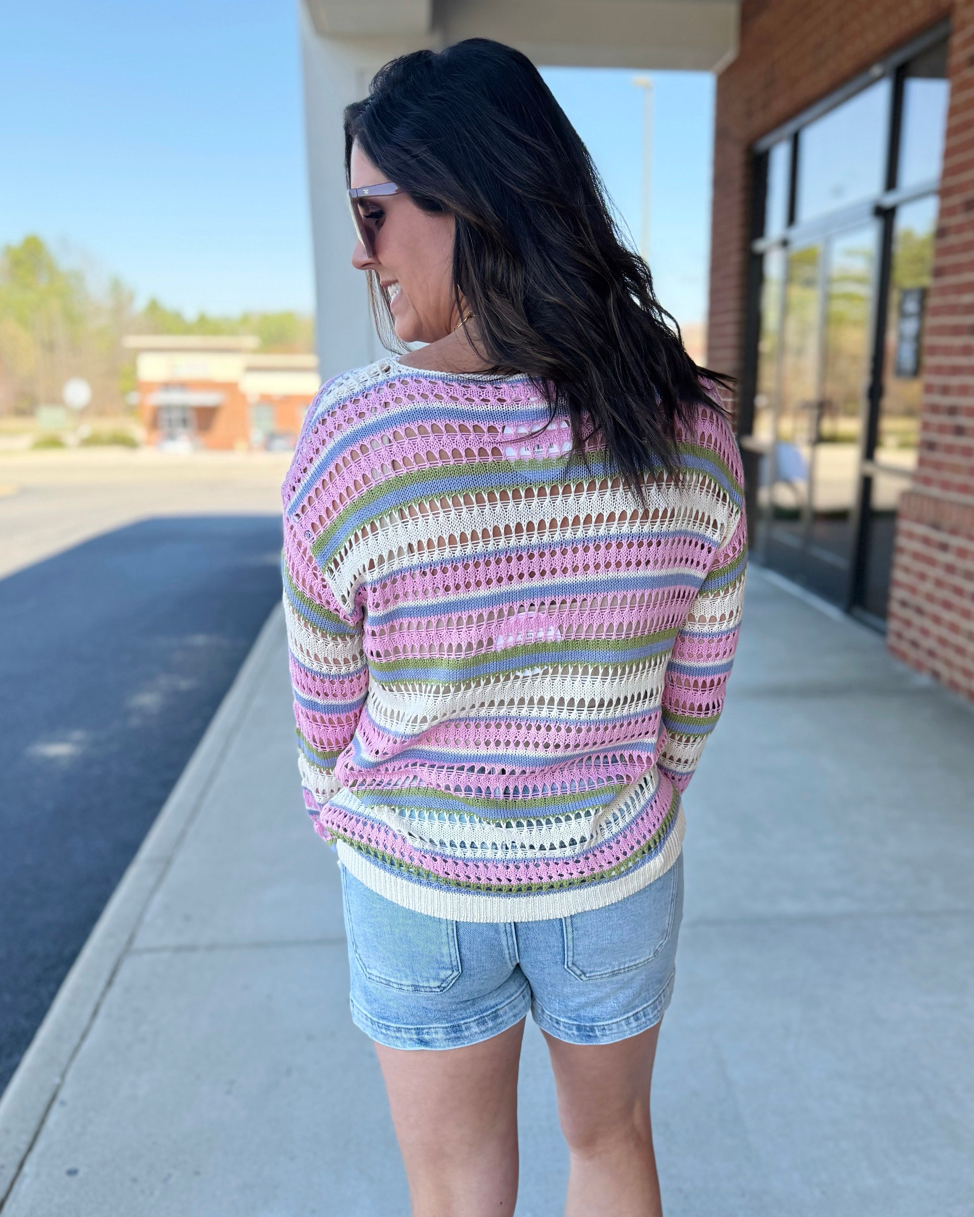 Woman wearing a colorful striped sweater and denim shorts standing on a sidewalk.