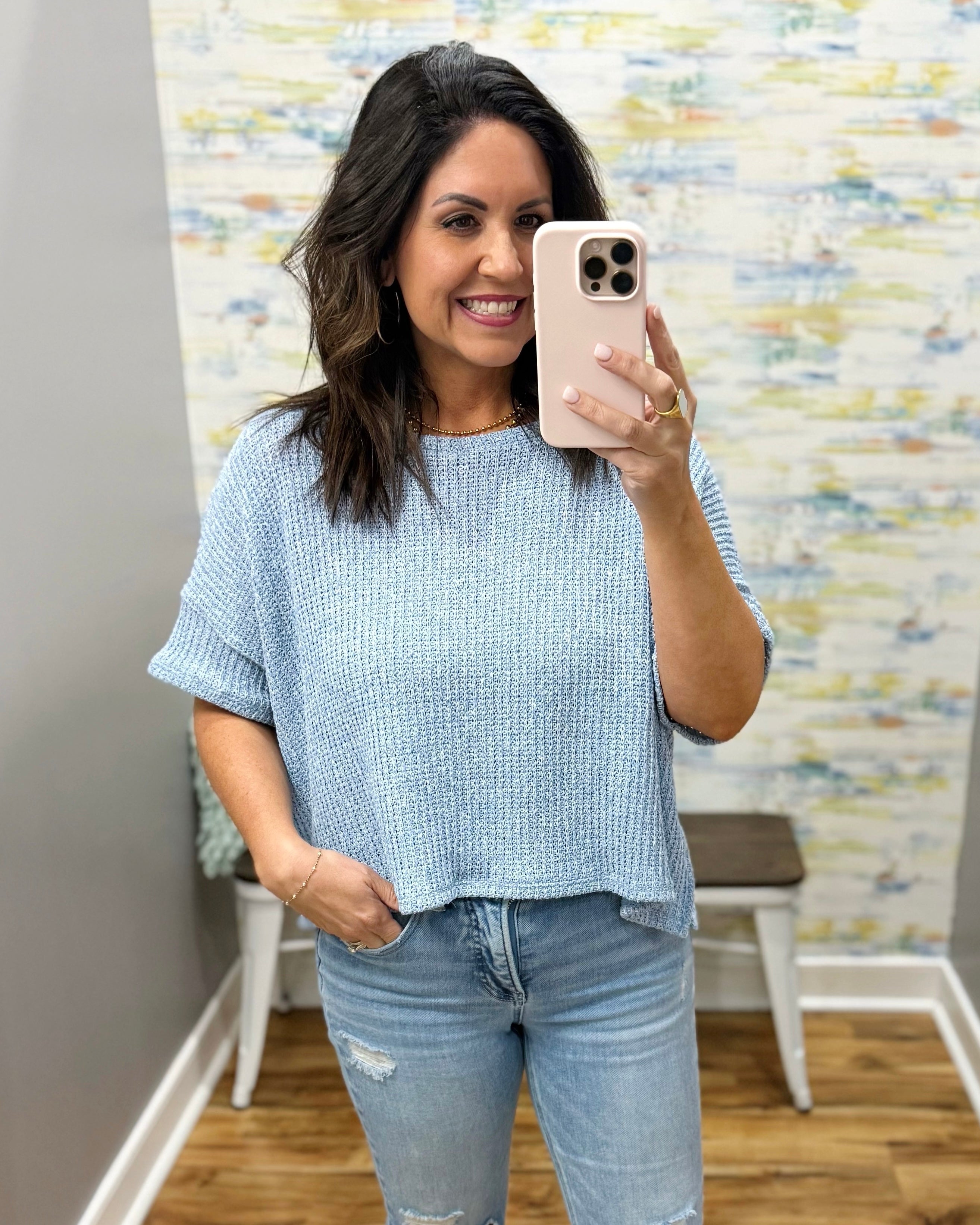 Woman taking a mirror selfie wearing a light blue sweater and jeans in a room with a textured wall.