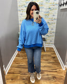 A woman takes a mirror selfie wearing a bright blue crewneck sweatshirt, blue jeans, and beige sneakers in a home hallway.