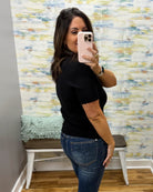 A woman takes a mirror selfie wearing a black short-sleeve knit top with a scalloped hem and blue jeans, standing in front of a colorful abstract wallpapered wall.