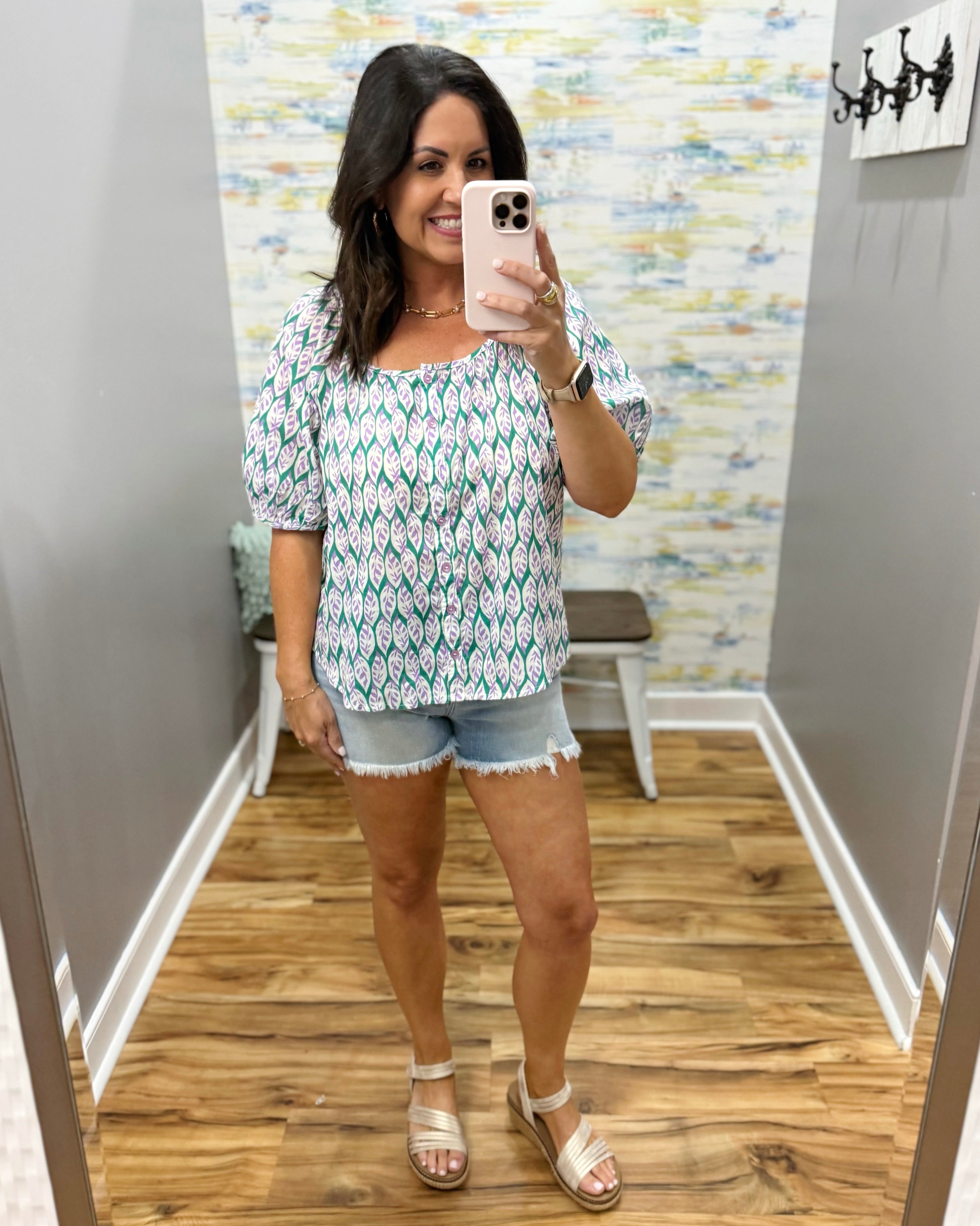 Woman taking a mirror selfie wearing a patterned top and denim shorts in a room with wooden flooring and a colorful wall.