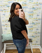 A woman taking a mirror selfie in a fitting room, wearing a black textured short-sleeve top and blue jeans.