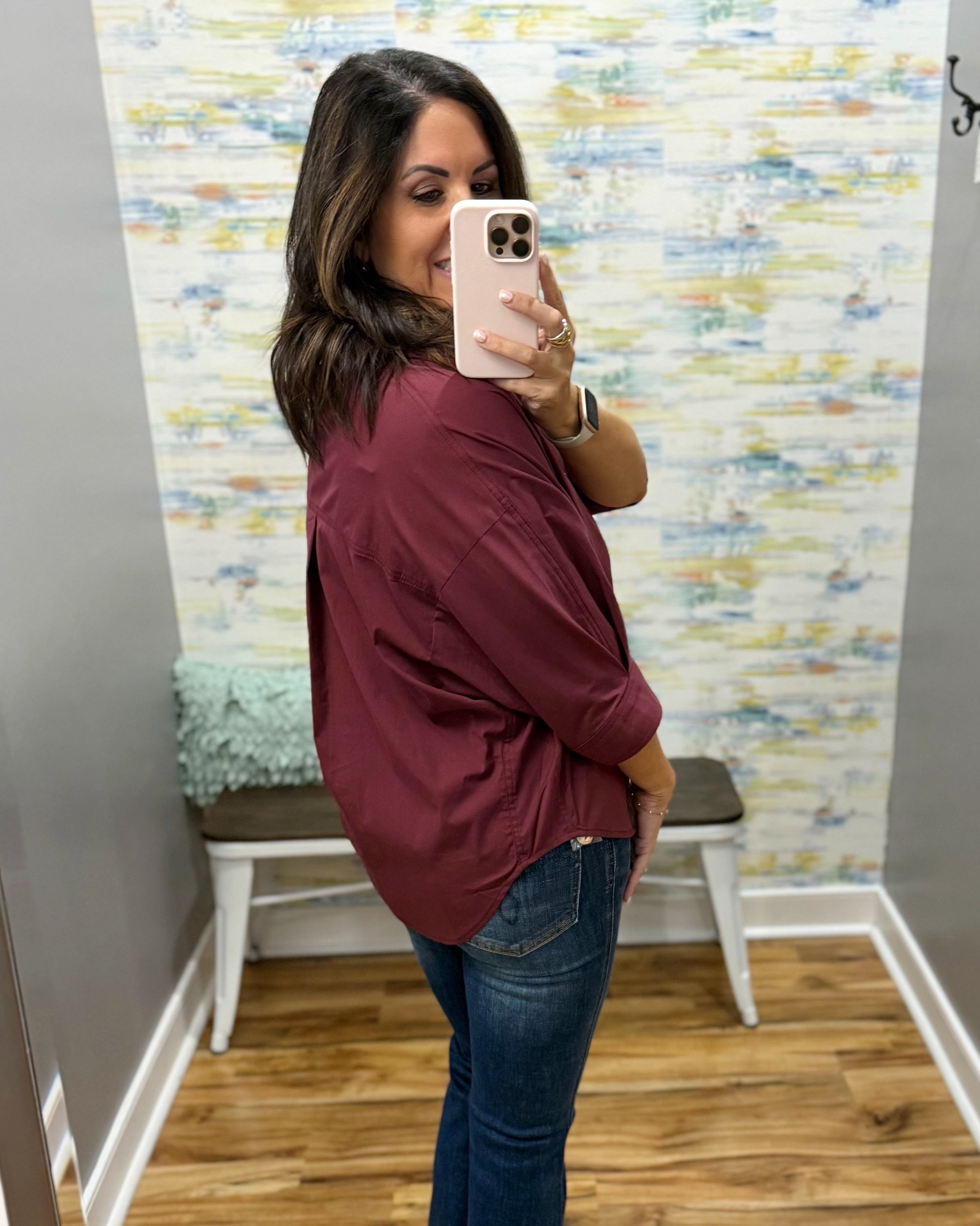 A woman takes a mirror selfie wearing a loose-fitting, long-sleeve burgundy button-up shirt and blue jeans in a fitting room with a patterned wallpaper background.