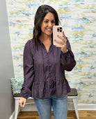 A woman takes a mirror selfie wearing a dark purple long-sleeve blouse with a small white floral pattern, paired with blue jeans. The background features a colorful abstract patterned wall and a white bench.