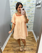 Woman taking a mirror selfie wearing a peach dress with red trim in a room with a patterned wall.