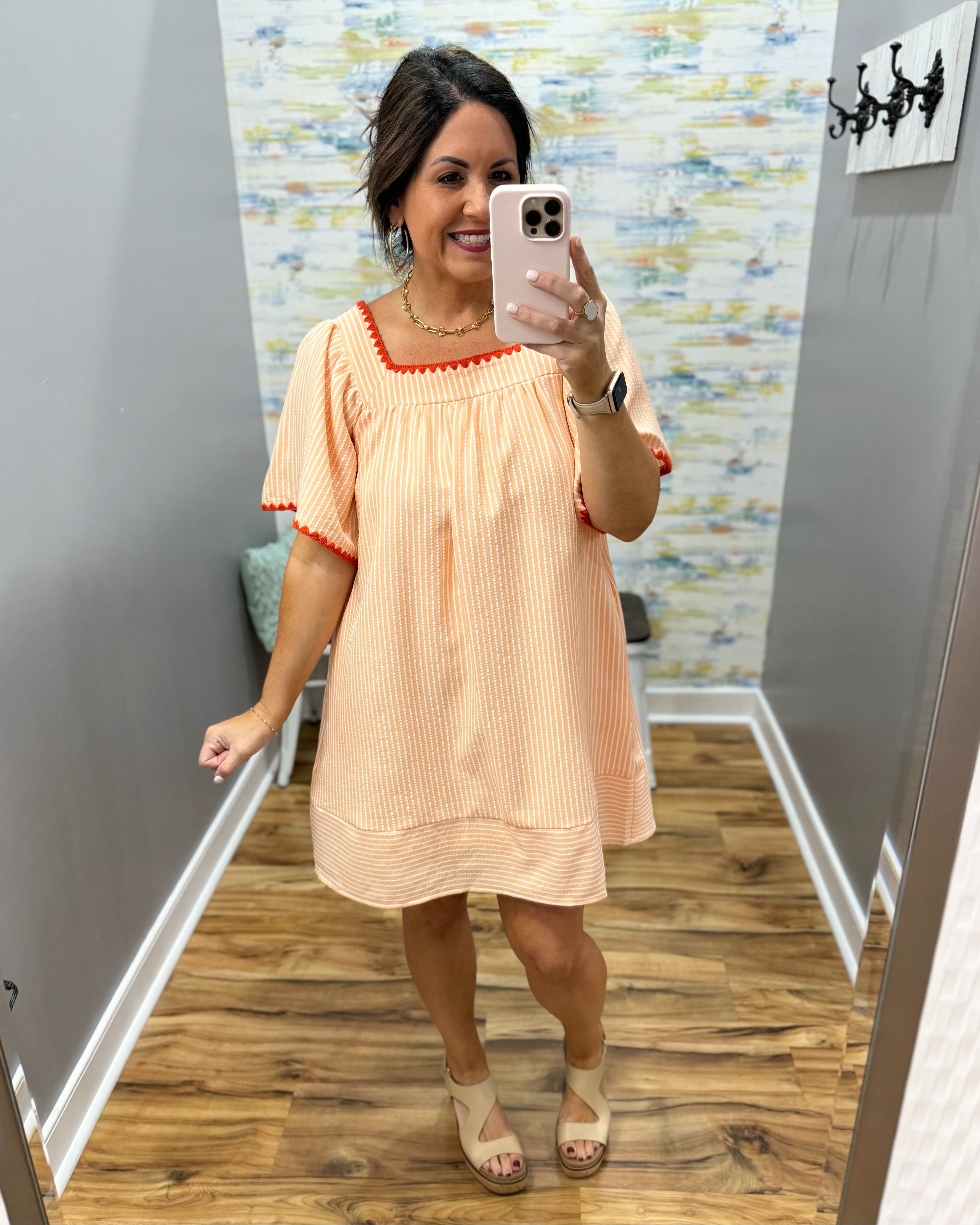 Woman taking a mirror selfie wearing a peach dress with red trim in a room with a patterned wall.