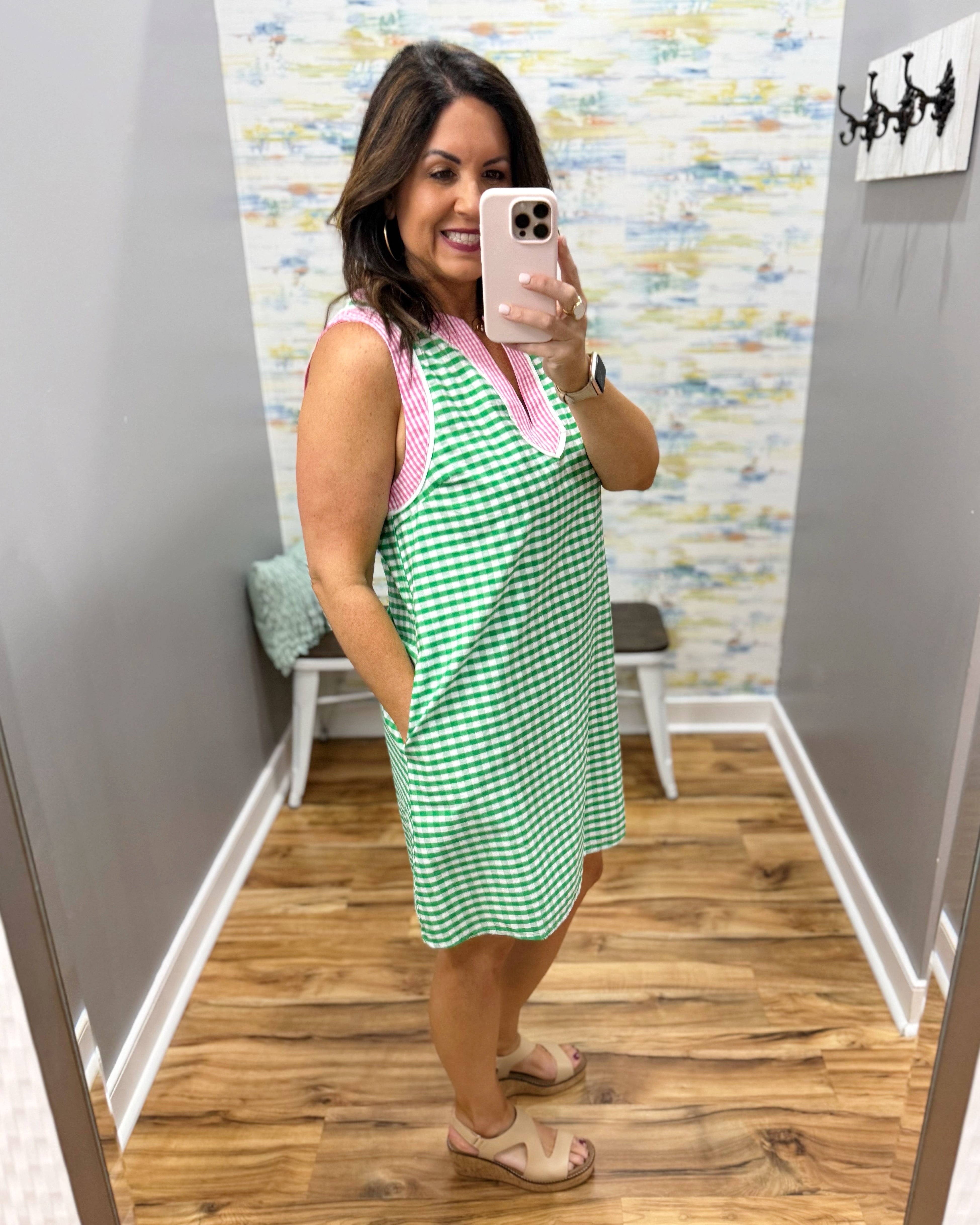 Woman taking a mirror selfie wearing a green and pink checkered dress in a room with a patterned wall.