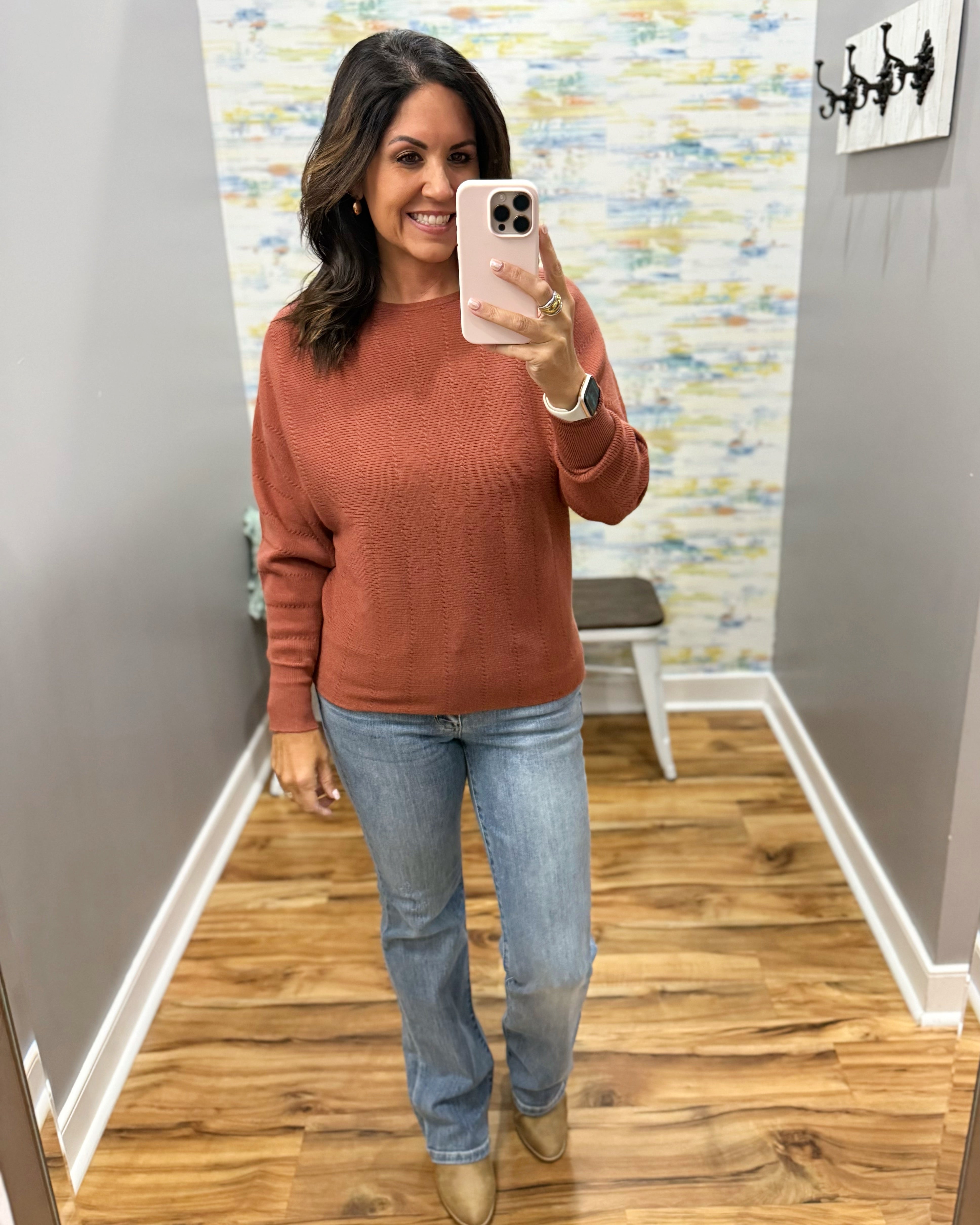 A woman takes a mirror selfie wearing a rust-colored long-sleeve knit sweater, light-wash blue jeans, and tan ankle boots in a home entryway.
