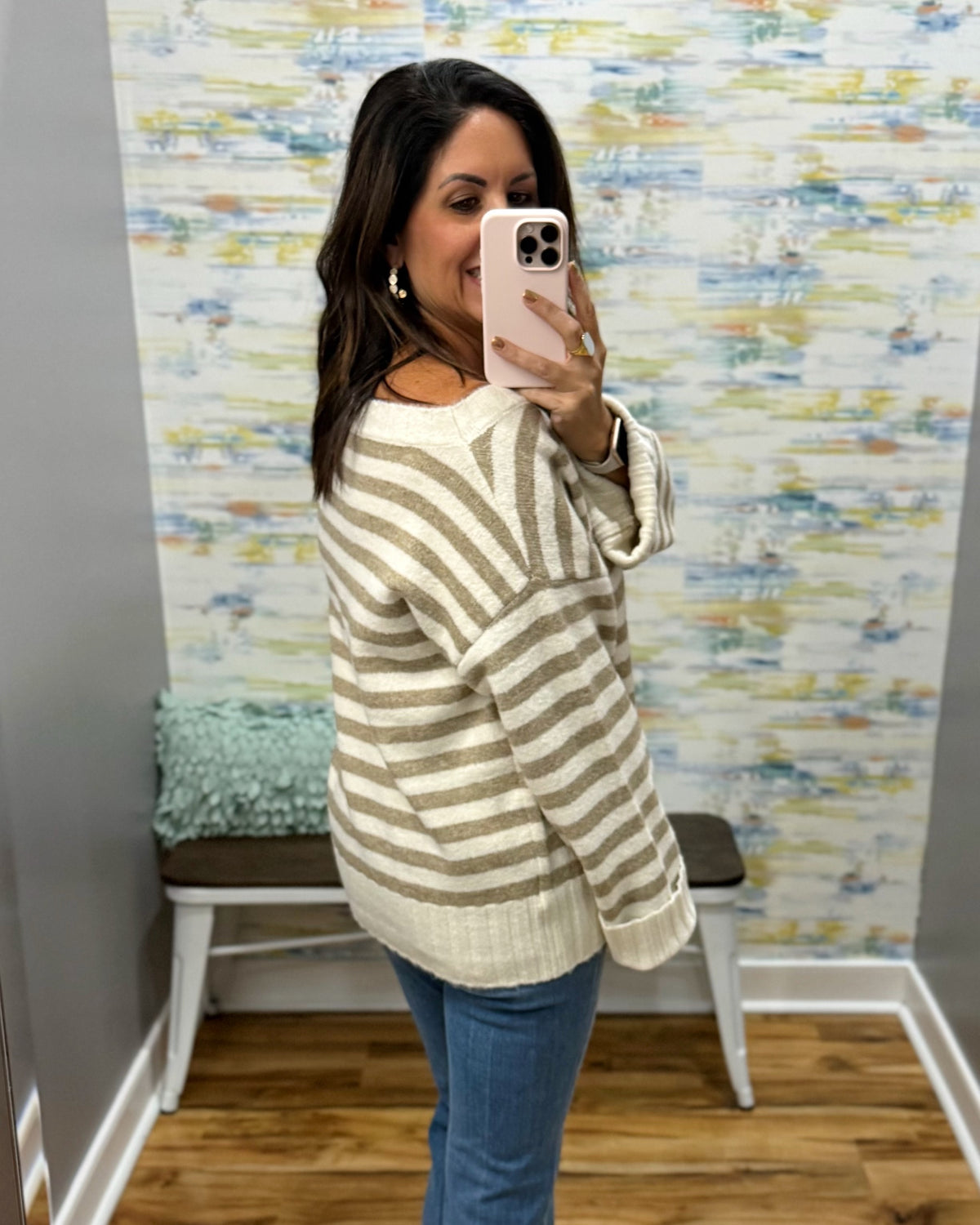 A woman takes a mirror selfie wearing a cream and taupe striped knit sweater, blue jeans, and a light pink phone. The background shows a patterned wallpaper, a wooden floor, and a small bench.