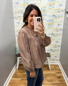 A woman takes a mirror selfie wearing a taupe brown long-sleeve blouse with gathered detailing and blue jeans in a fitting room with wood floors and a patterned wall.