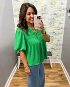 Woman taking a mirror selfie wearing a green top and blue jeans in a room with a colorful wall.