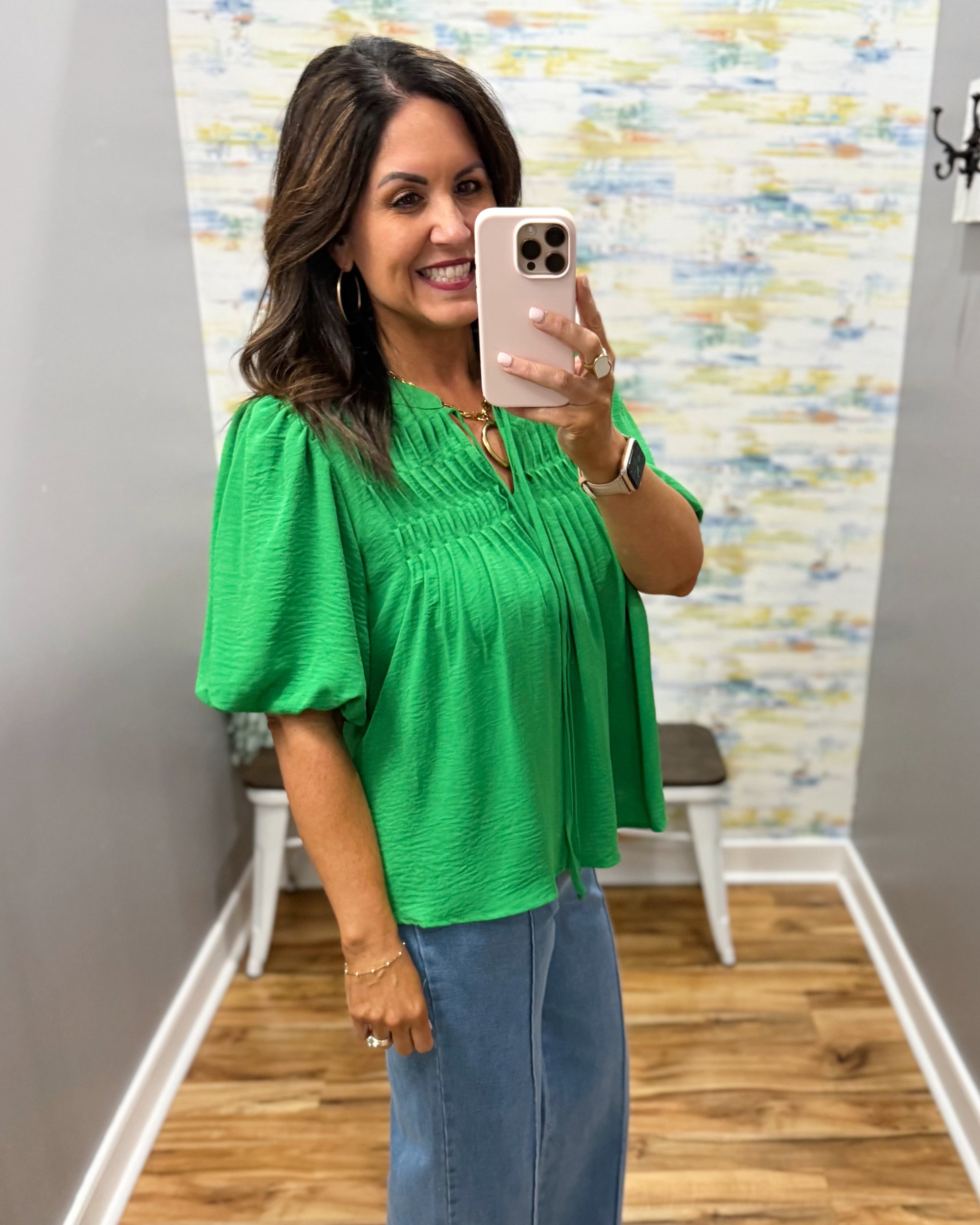 Woman taking a mirror selfie wearing a green top and blue jeans in a room with a colorful wall.