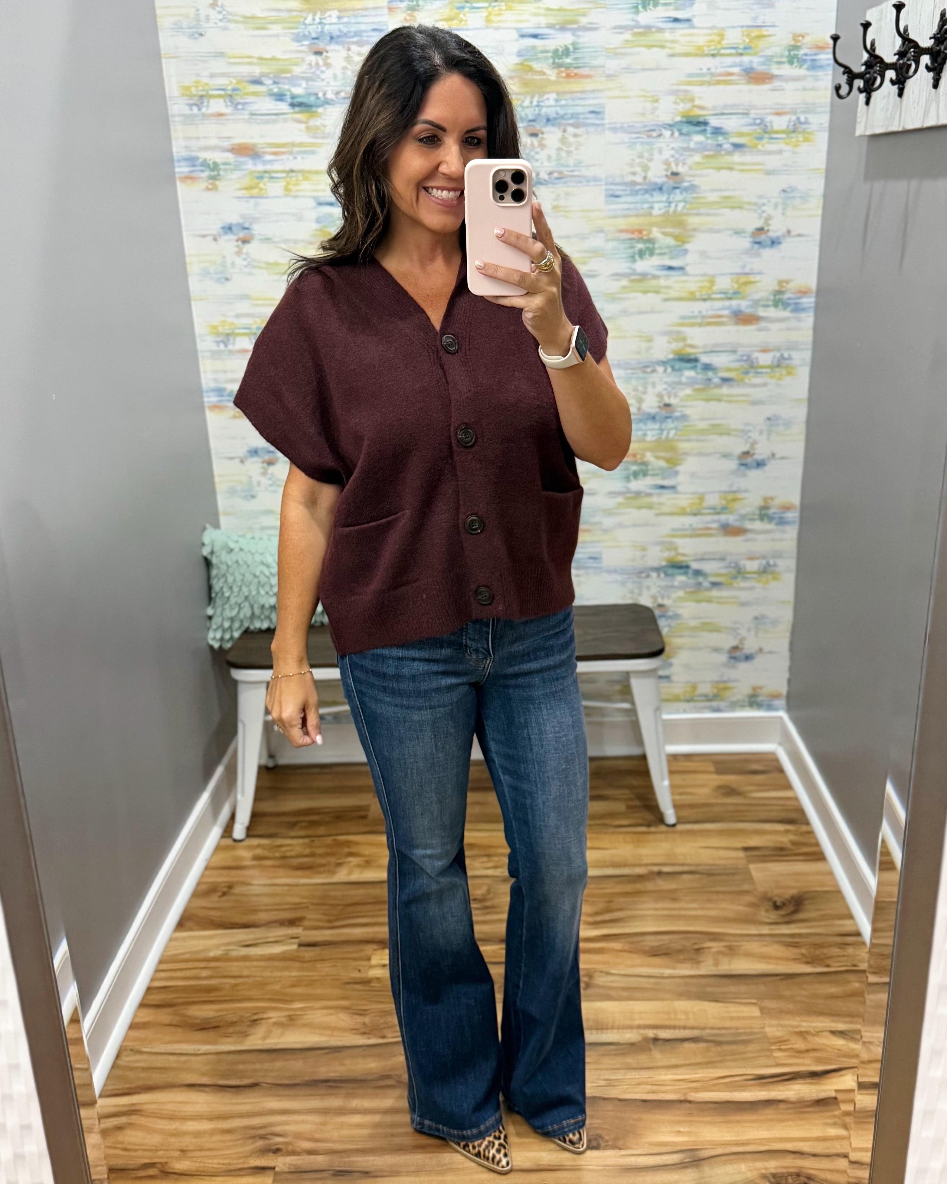 A woman takes a mirror selfie in a fitting room wearing a short-sleeve button-front top in deep burgundy and blue flare jeans.