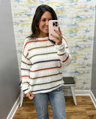 A woman takes a mirror selfie wearing a cream knit sweater with horizontal stripes in rust orange, olive green, and navy blue, paired with blue jeans.