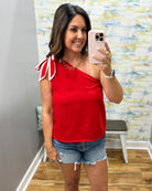Woman taking a mirror selfie wearing a red one-shoulder top and denim shorts.