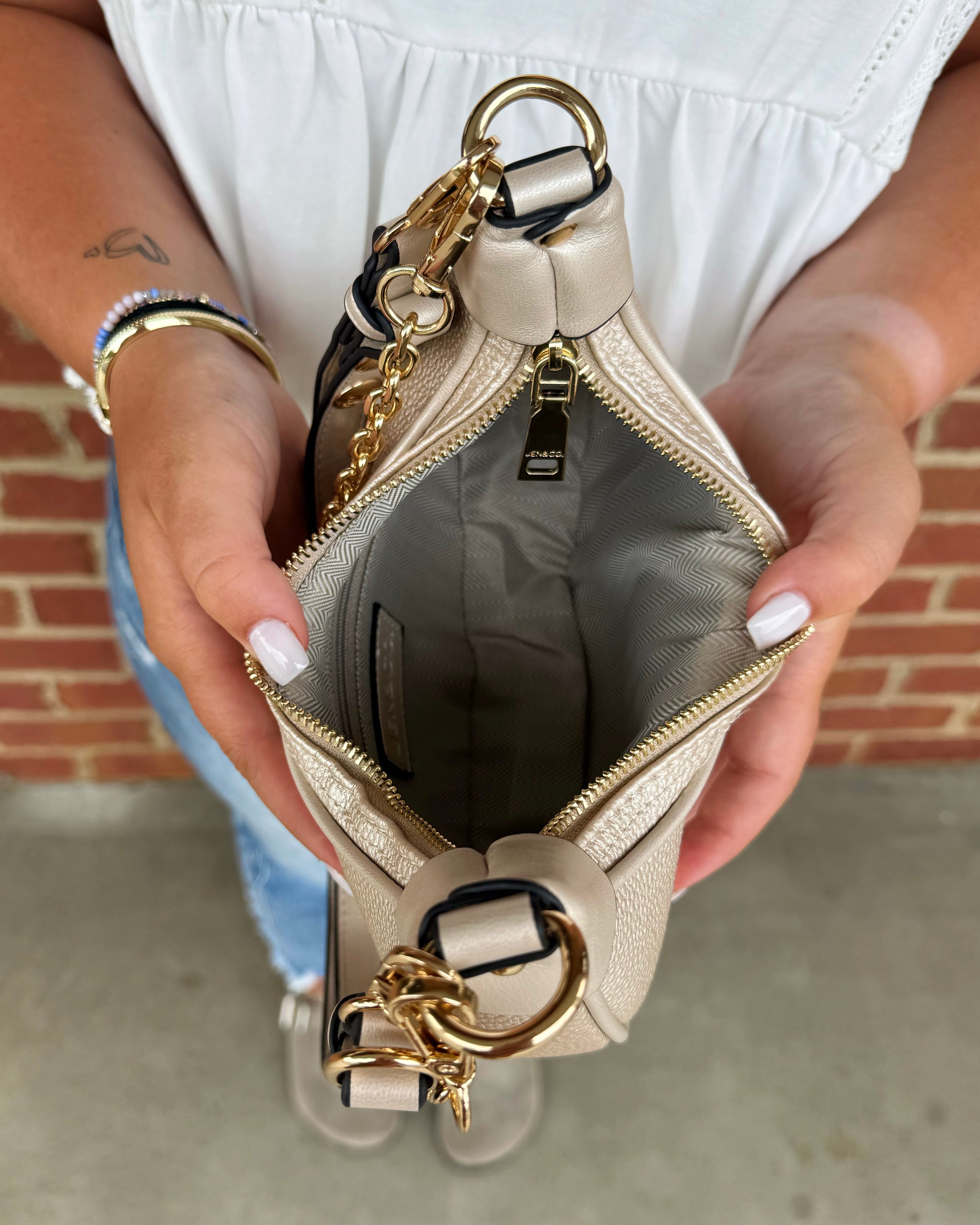 A person holds open a small beige leather clutch or wallet-style handbag to show the interior. The bag has gold-tone hardware, a chain strap, and a ring detail, with a gray interior lining.