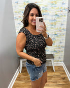 Woman taking a mirror selfie wearing a black floral top and denim shorts in a room with colorful wall art.