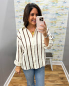 A woman takes a mirror selfie wearing a cream and brown vertically striped waffle-knit button-down shirt and blue jeans in a fitting room.